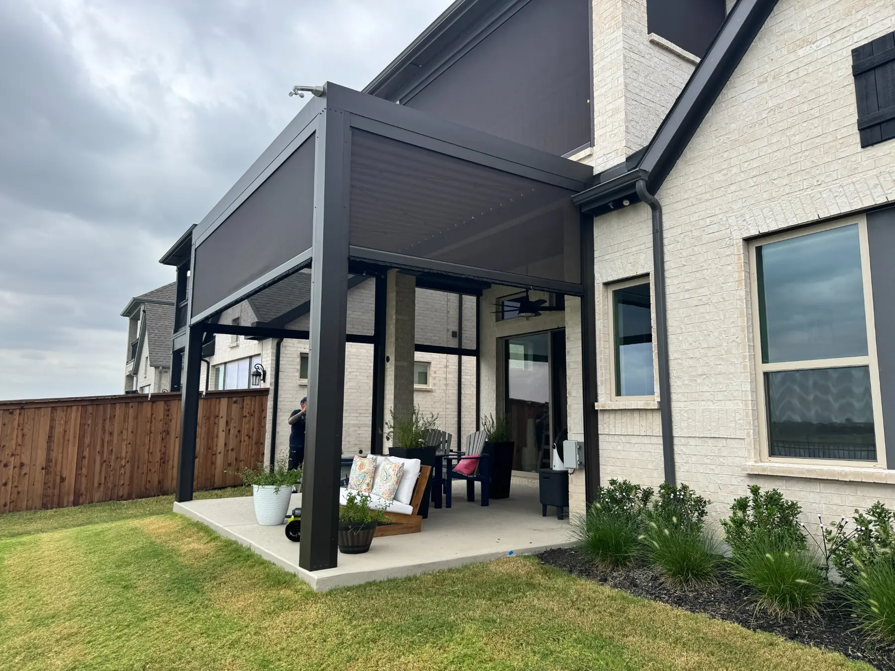 Motorized patio screens partially lowered around a black louvered roof attached to a white brick home.