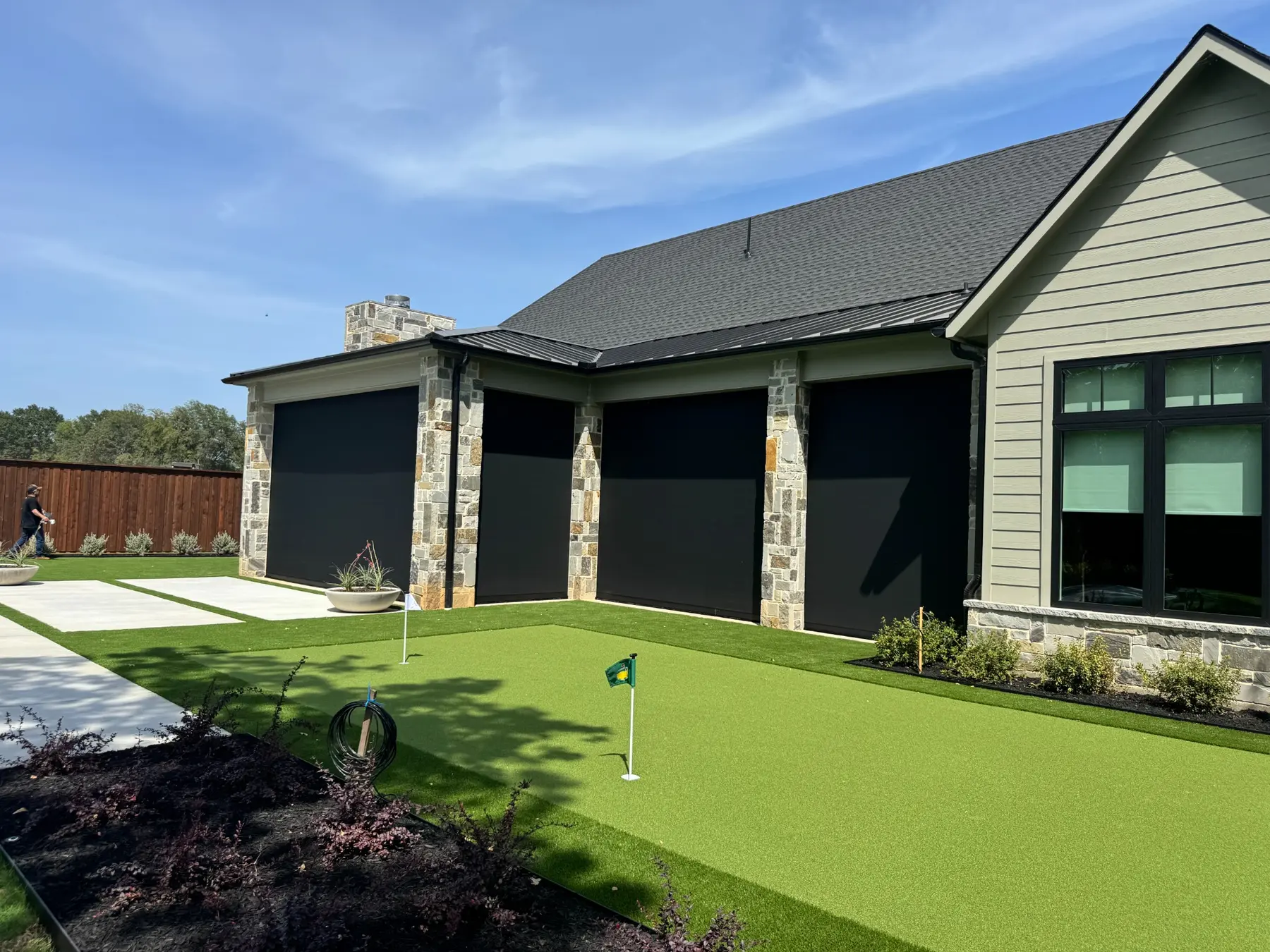 Three large black motorized patio screens on a stone home beside a backyard putting green.